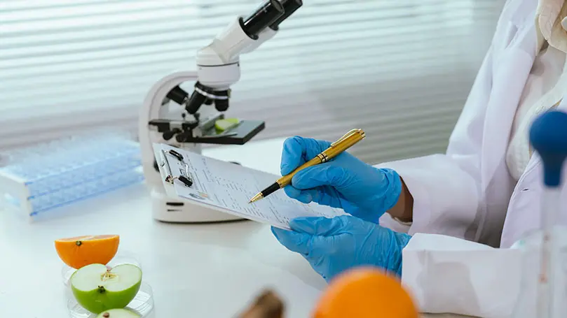 Labortisch mit Mikroskop, im Fokus zwei Hände die ein Klemmbrett halten