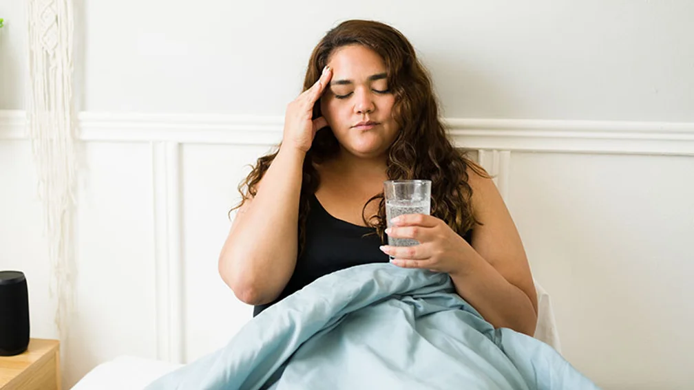 junge dunkelhaarige Frau sitzt mit Bettdecke über dem Schoß auf dem Bett, ein Glas Sprudelwasser in der Hand, die andere Hand hält sie an die schmerzende Schläfe
