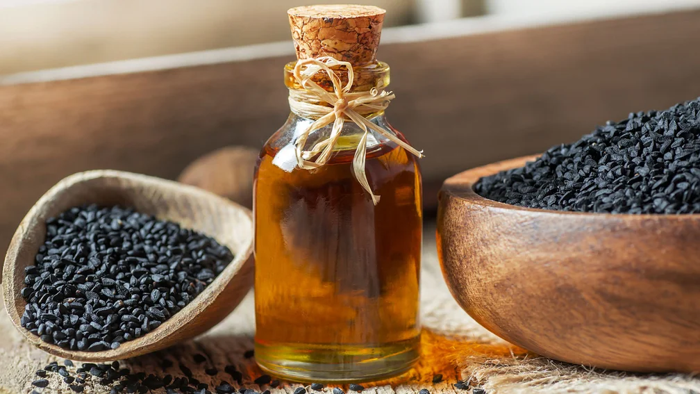 Glasflasche gefüllt mit Schwarzkümmelöl, daneben liegt ein Holzlöffel und eine Holzschale jeweils mit Samen befüllt