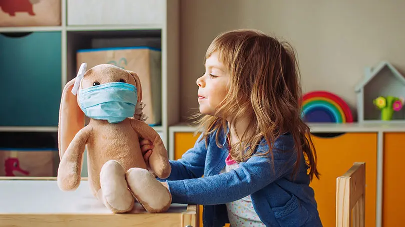 Mädchen im Kindergarten betrachtet Stofftier-Hund, das eine OP-Maske trägt