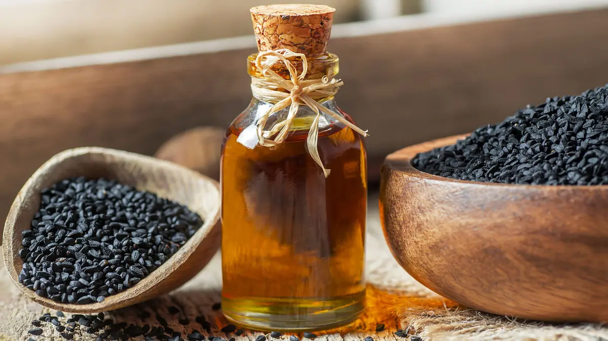 Glasflasche gefüllt mit Schwarzkümmelöl, daneben liegt ein Holzlöffel und eine Holzschale jeweils mit Samen befüllt