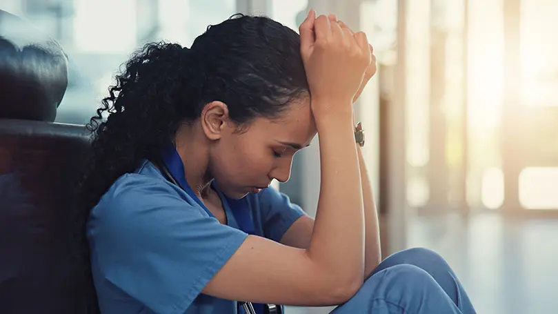 junge Krankenschwester sitzt mit angezogenen Knien auf dem Boden, den Kopf auf die Arme gestützt