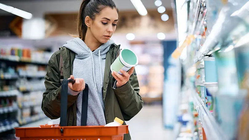 Junge Frau im Supermarkt mit Einkaufskorb in der Hand betrachtet einen Joghurtbecher