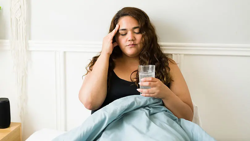 junge dunkelhaarige Frau sitzt mit Bettdecke über dem Schoß auf dem Bett, ein Glas Sprudelwasser in der Hand, die andere Hand hält sie an die schmerzende Schläfe