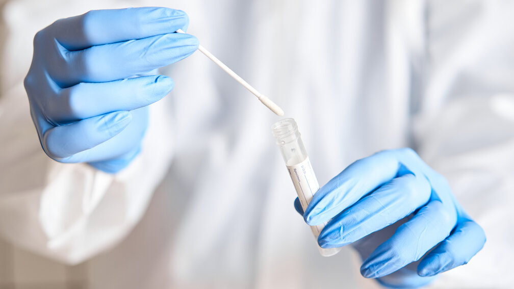 Hände in blauen Handschuhen halten Coronateststäbchen und Röhrchen