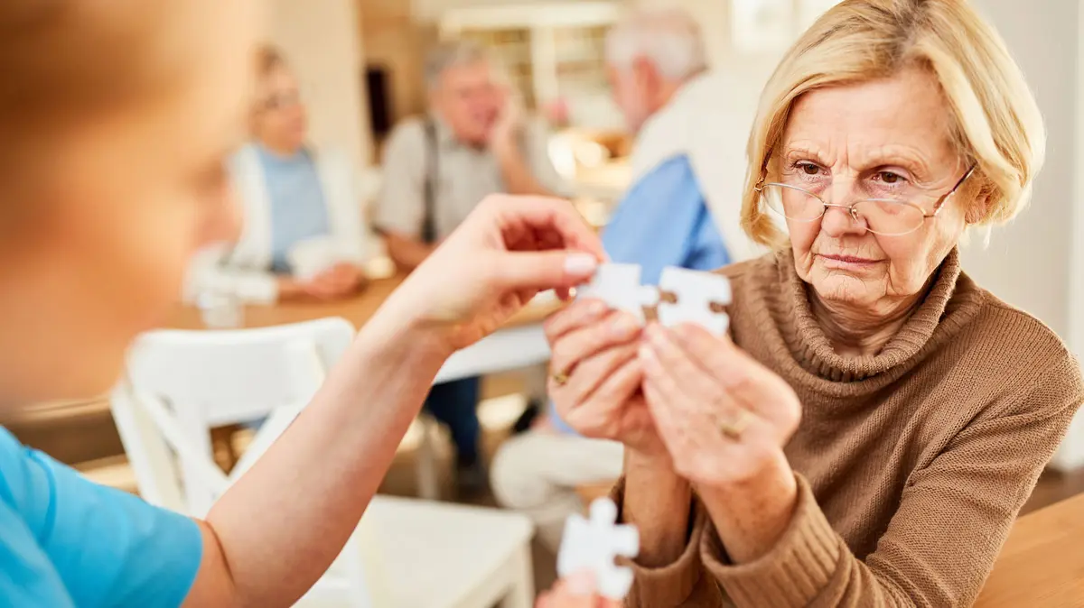 Seniorin im Altersheim und hält Puzzelteile in den Händen, gegenüber sitzt eine Pflegerin 