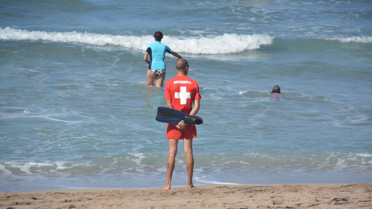 Rettungsschwimmer steht am Rand des Gewässers; zwei Leute befinden sich im Wasser