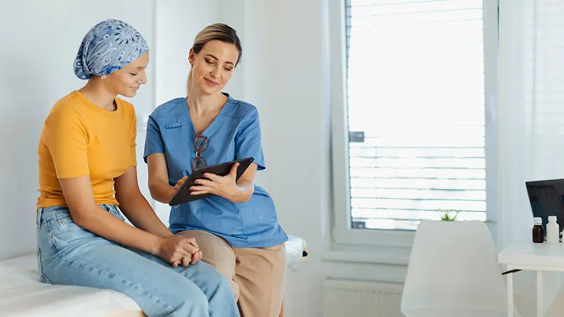 junge Krebspatienten sitzt auf Krankenhausliege, MFA zeigt ihr etwas auf einem Tablet