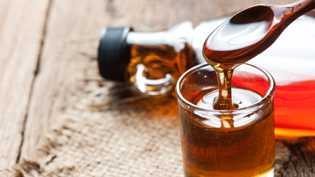 Ein mit Ahornsirup gefülltes Glas mit Holzlöffel, von dem Sirup ins Glas tropft