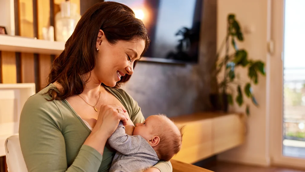 Fläschchen mit Muttermilch, im Hintergrund stillt die Mutter ihren Säugling an der Brust