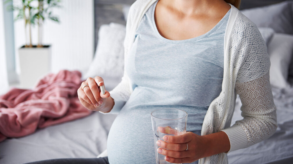 Schwangere Frau hält Tablette in der einen Hand und in der anderen ein Glas Wasser