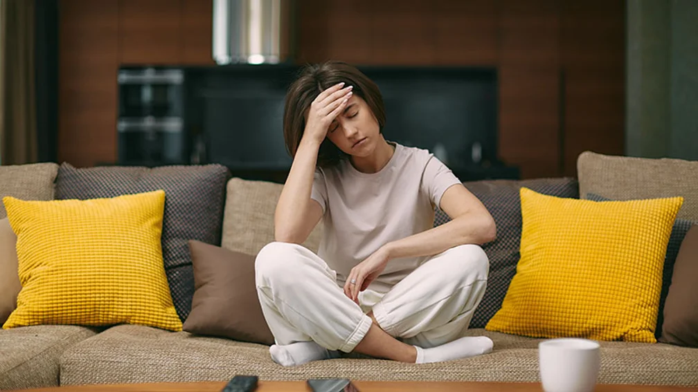 junge Frau sitzt im Schneidersitz auf einem Sofa und stützt erschöpft ihren Kopf in die rechte Hand