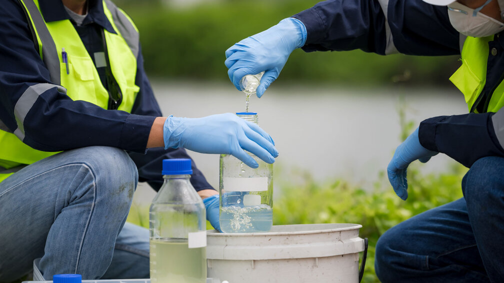 zwei Männer nehmen mithilfe von Gefäßen Proben vom Abwasser
