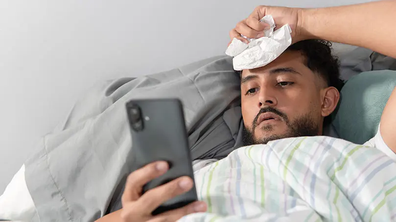 Mann mit Fieber im Bett schaut auf sein Smartphone
