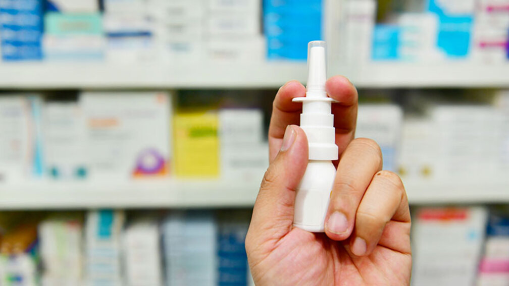 männliche Hand hält Nasenspray vor Apothekenregal