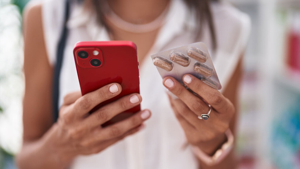 Frau hält Handy in der einen hand und einen Tablettenblister in der anderen Hand