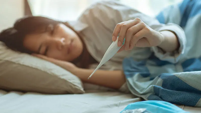 junge Asiatin liegt im Bett und schaut auf ein Fieberthermometer