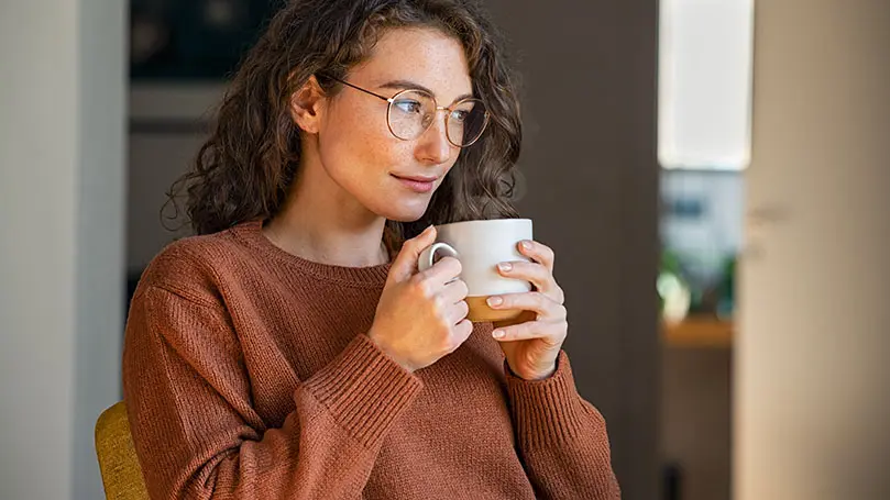 junge, braunhaarige Frau mit Brille hält weiße Keramiktasse mit Tee oder Kaffee in der Hand