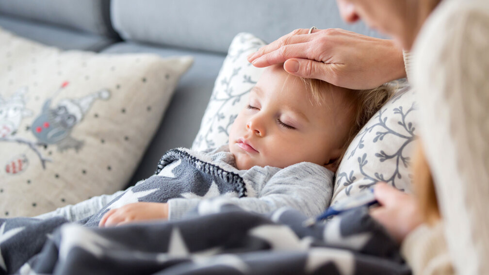 Kleinkind liegt kränkelnd auf dem Sofa, Hand der Mutter fasst die Stirn an