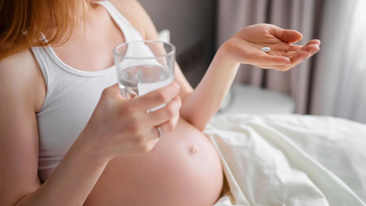 Schwangere liegt im Bett und hält eine Tablette in der einen Hand und ein Glas Wasser in der anderen