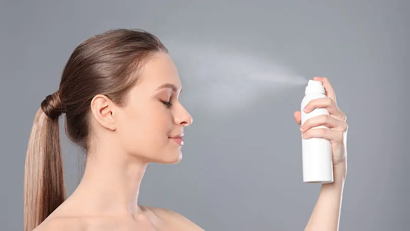 Langhaarige Frau sprüht sich mit geschlossenen Augen Spray ins Gesicht