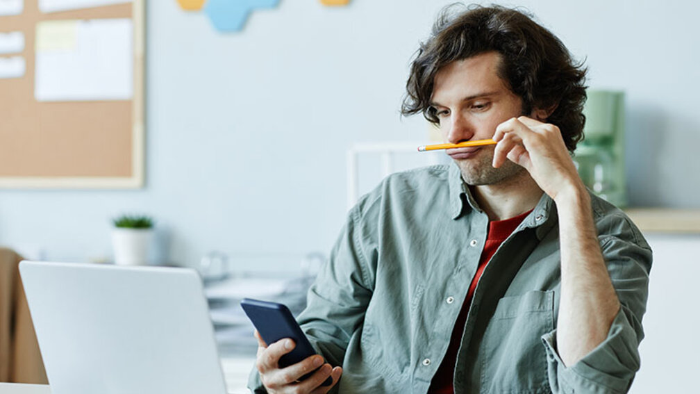 Junger Mann sitzt vor Laptop, schaut aufs Handy und hält Stift unter die Nase