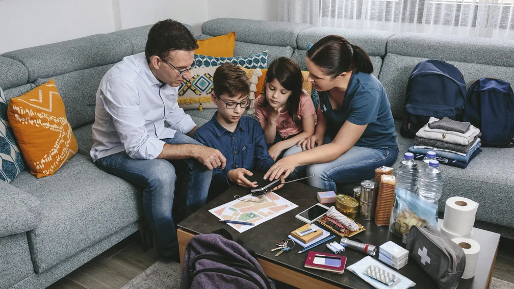 Familie sitzt auf dem Sofa, vor ihnen diverse Gegenstände wie Taschenlampe, Verbandskasten, Wasser, Batterien, usw.