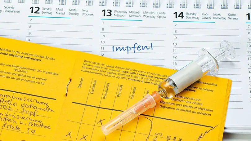 Eine Impfsspritze und ein aufgeschlagenes Impfbuch liegen auf einem Kalender
