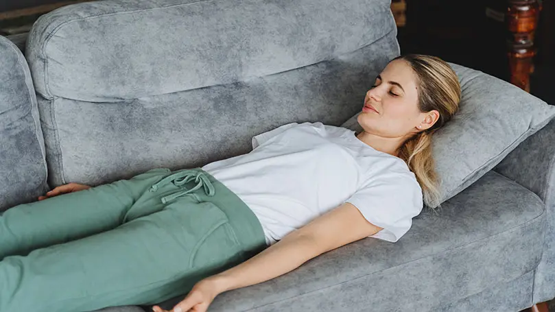 blonde junge Frau liegt mit geschlossenen Augen entspannt auf einem grauen Sofa