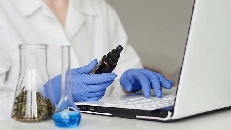 Frau mit Pipettenflasche in Hand recherchiert mit Handschuhen am Laptop