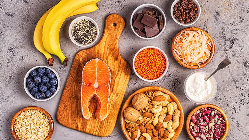 Bananen, Nüsse, Blaubeeren, dunkle Schokolade, Haferflocken und rote Linsen in weißen Schälchen auf Steinuntergrund