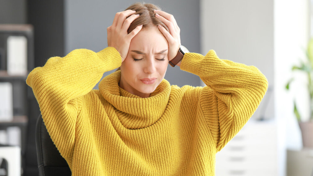 Eine  junge Frau in einem gelben Pullover hält ihren Kopf mit beiden Händen. Ihr Gesichtsausdruck deutet auf starke Kopfschmerzen hin.