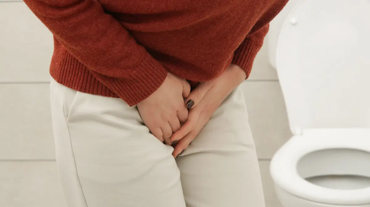 Frau steht seitlich vor einer Toilette und fasst sich mit beiden Händen in den Schritt