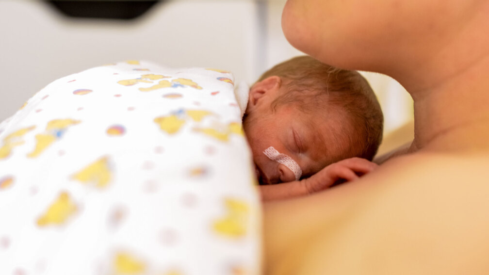 Frühgeborenes liegt auf der Brust der Mutte in eine Decke eingewickelt