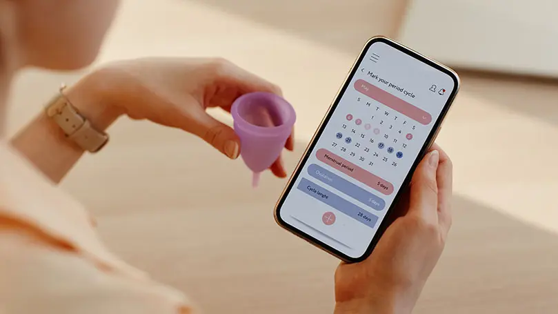 Frau hält in linker Hand Menstruationstasse, in rechter Hand ein Smartphone, auf dem eine Zyklus-App geöffnet ist