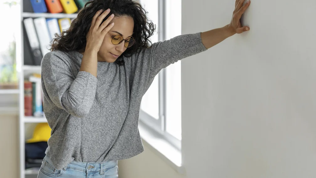 Frau hält sich stehend mit einer Hand an der Wand und die andere Hand an den Kopf