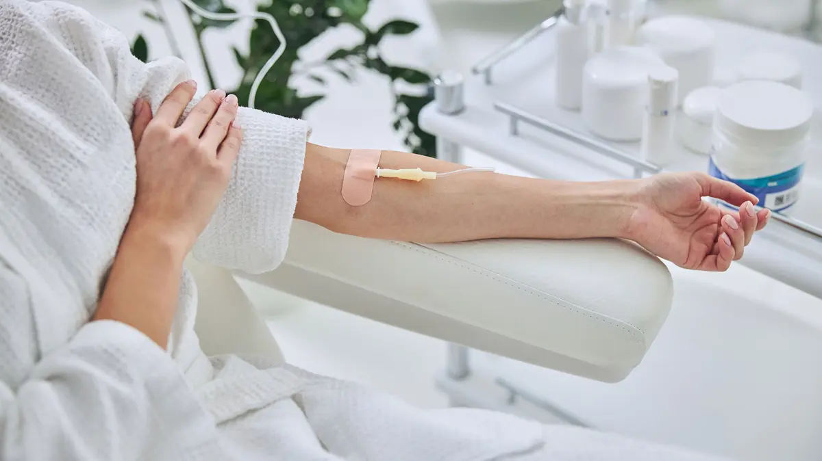 Frau im Bademantel auf einem Stuhl, mit einer Infusionsnadel in der Armbeuge
